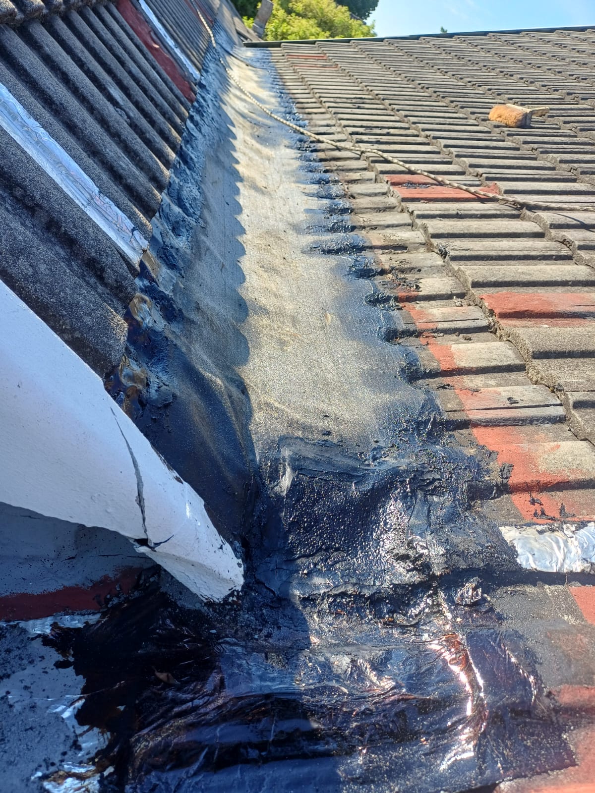 Close-up view of a tiled pitched roof valley sealed with a thick, black torch-on waterproofing membrane overlapping the edges of the roof tiles to prevent water leaks.