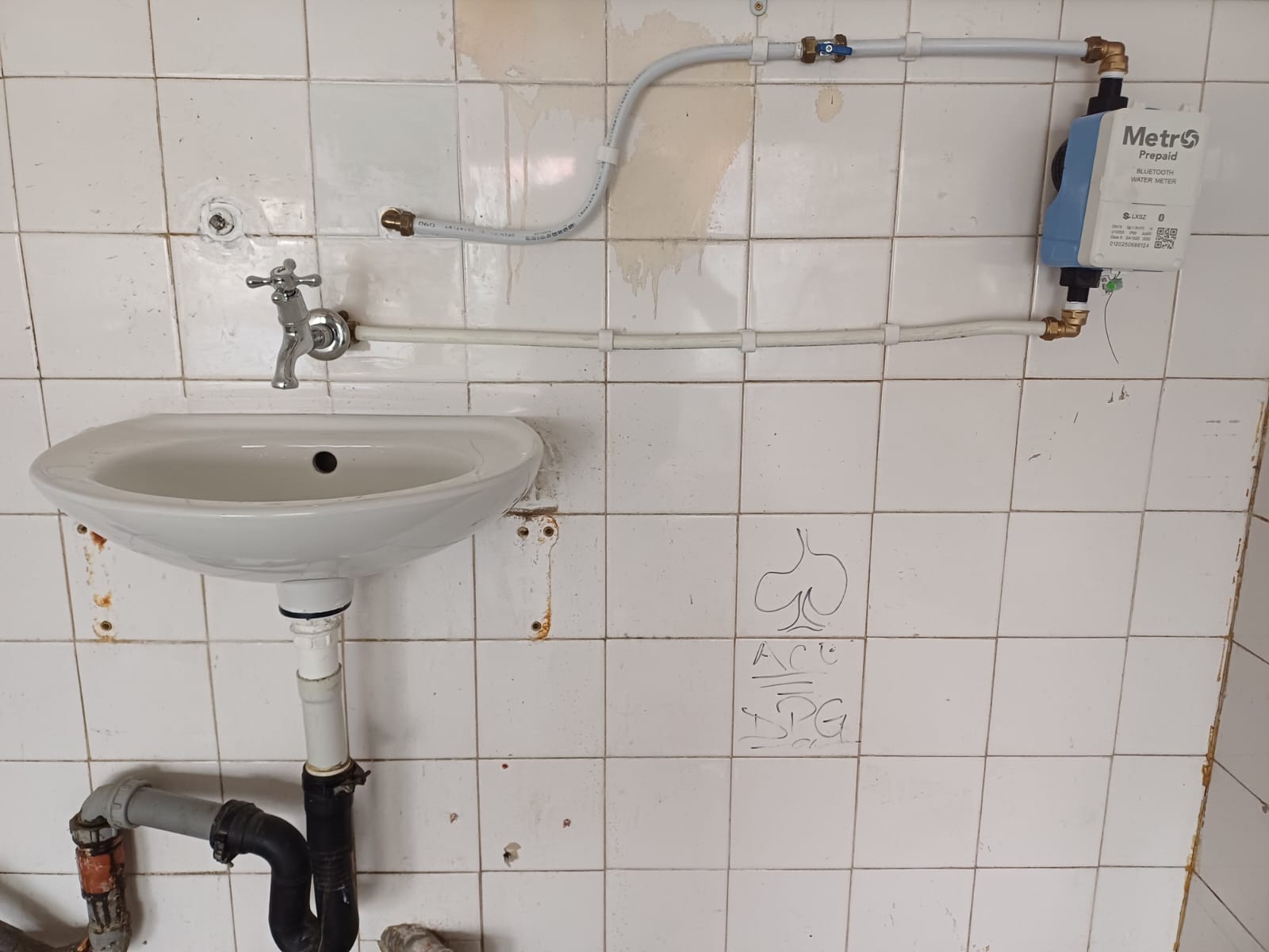 A wall-mounted ceramic wash basin with surface-level white plumbing pipes connecting to a blue and white Metro Prepaid water meter installed on a white tiled wall.