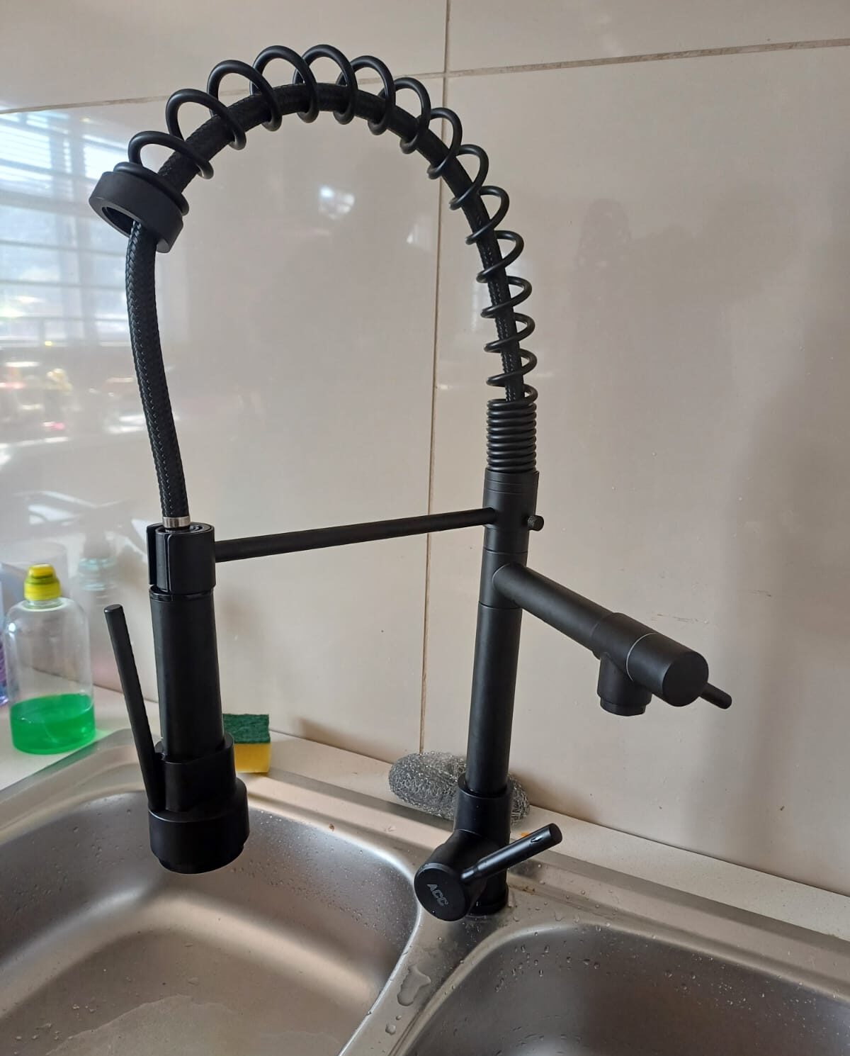 A newly installed matte black commercial-style kitchen mixer tap featuring a coiled spring neck and a pull-down sprayer, mounted on a stainless steel double sink against a light tiled backsplash.