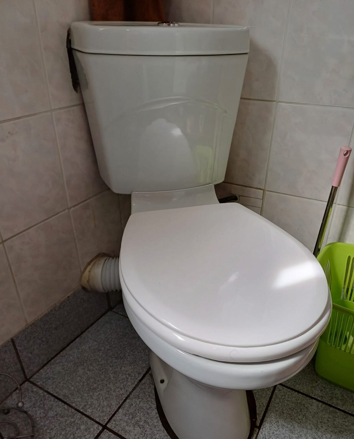 A newly installed white ceramic close-coupled toilet suite with a closed lid, fitted in a corner against light-colored wall tiles and grey floor tiles, showing a flexible pan connector at the rear.